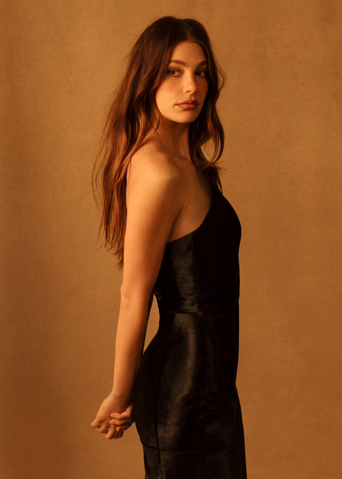 Camila Morrone wears a black dress in front of a gold backdrop.