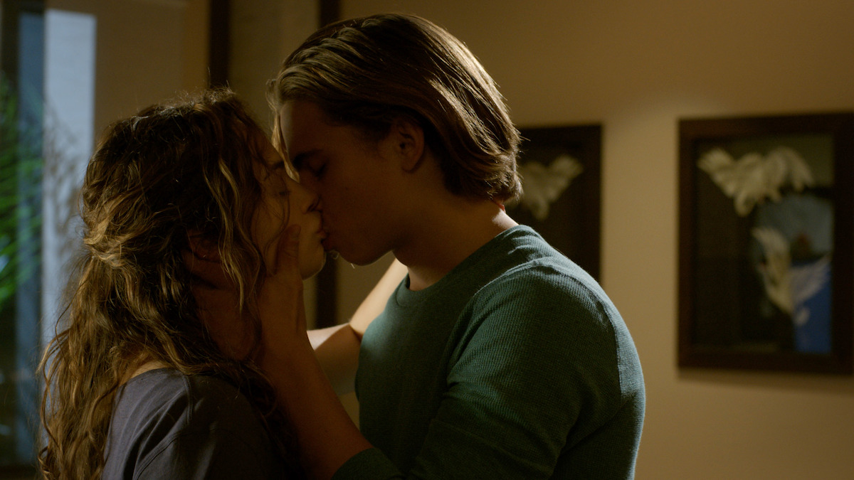 Two people share an intimate kiss in a warmly lit room with framed artwork and soft, ambient lighting in the background.