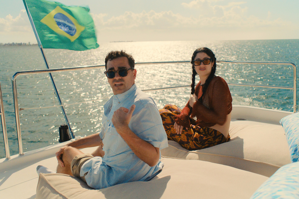 Two people relax on loungers on a yacht deck, wearing sunglasses, with a Brazilian flag waving behind them. The yacht is on the ocean near a coastal city under a sunny sky.