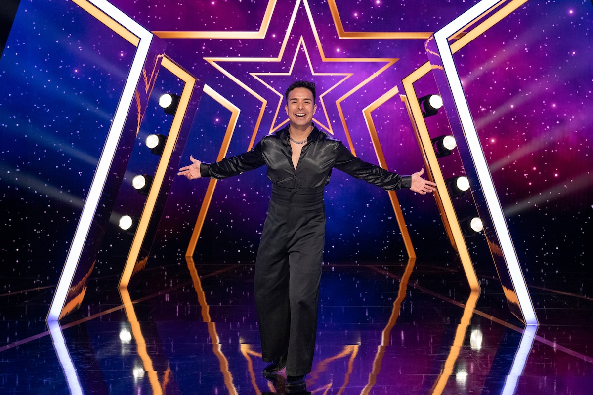 Fernando Velasco con un traje negro está de pie con los brazos abiertos en un escenario brillante, con una gran estrella iluminada y un fondo violeta vibrante con luces.