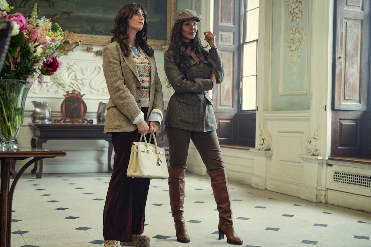 Two stylish women stand in an elegant, classic interior with tall windows and ornate decor. One holds a white handbag, both wear blazers and boots, appearing poised and fashionable in a well-lit, sophisticated room with flowers and art.