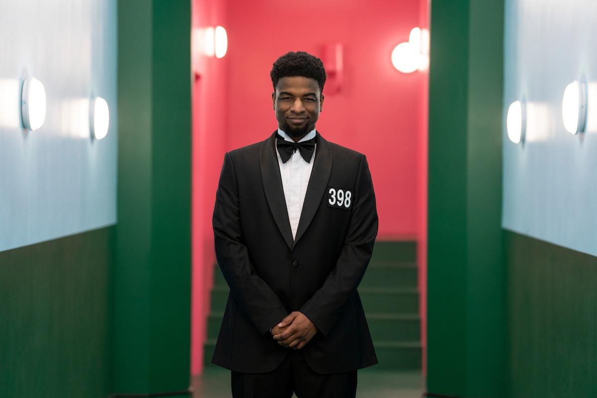 A man in a black tuxedo with the number 398 stands in a brightly lit, colorful hallway with green walls, light blue panels, and a red background, creating a formal and slightly tense atmosphere.