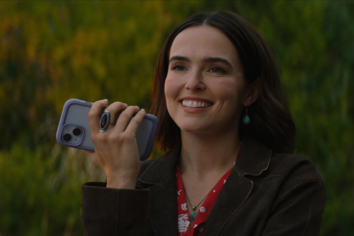 A smiling woman holding a smartphone outdoors, wearing a brown jacket and red shirt, standing in front of green trees and foliage in natural light.