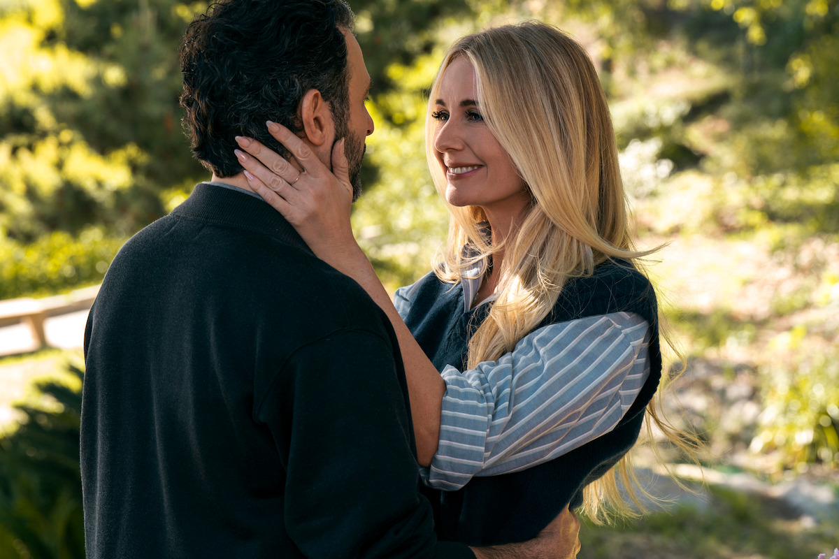 A woman and man stand close together outdoors in a sunny, green park. The woman smiles warmly, holding the man's face, creating a romantic and affectionate mood in a peaceful natural setting.