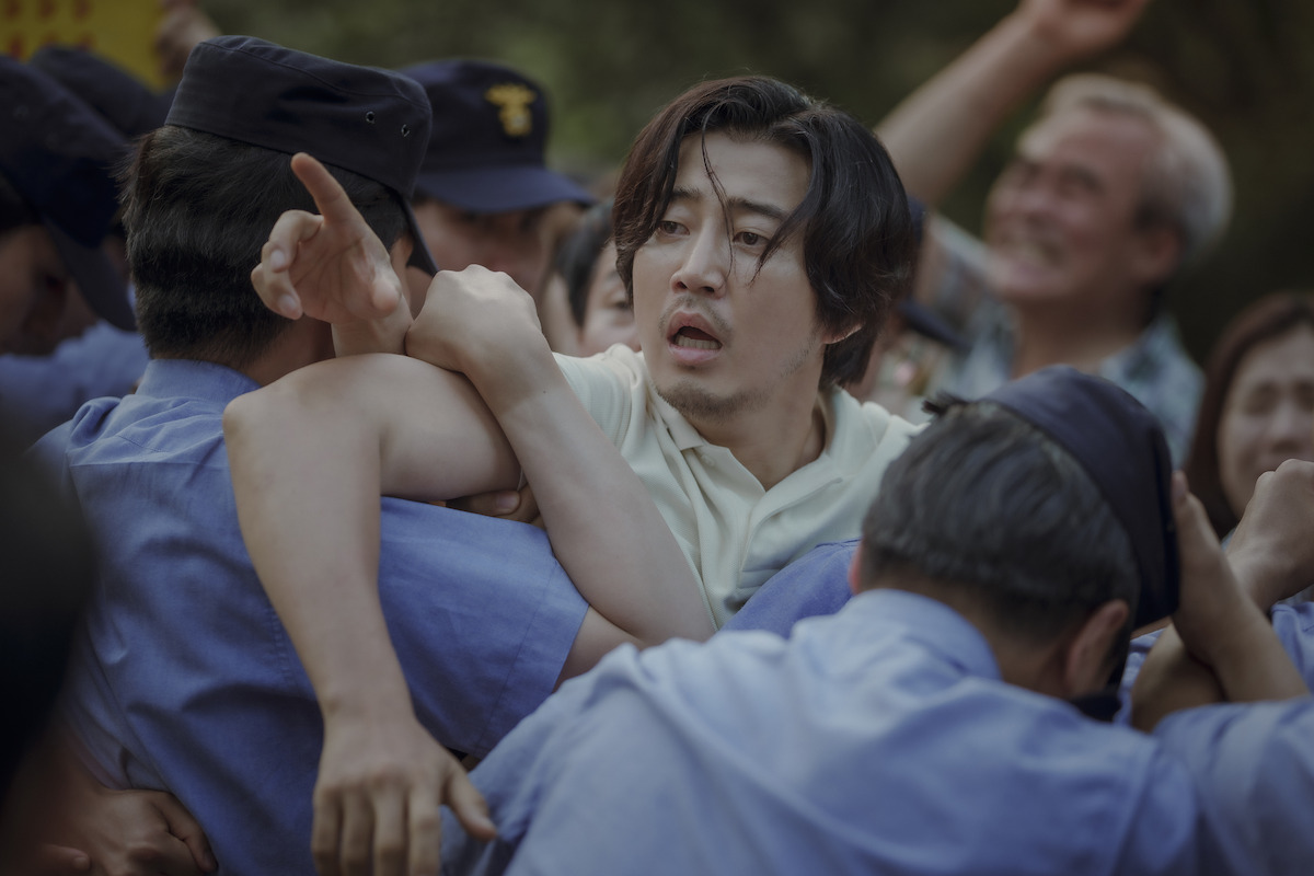 Yoon Kye-sang as Koo Sang-jun attempts with an exasperated look on his face attempts to run past police officers in a crowd of people in an image from the series ‘The Frog.’