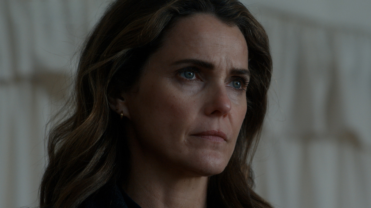 Woman with long brown hair and serious expression standing indoors in front of a softly lit beige curtain background.