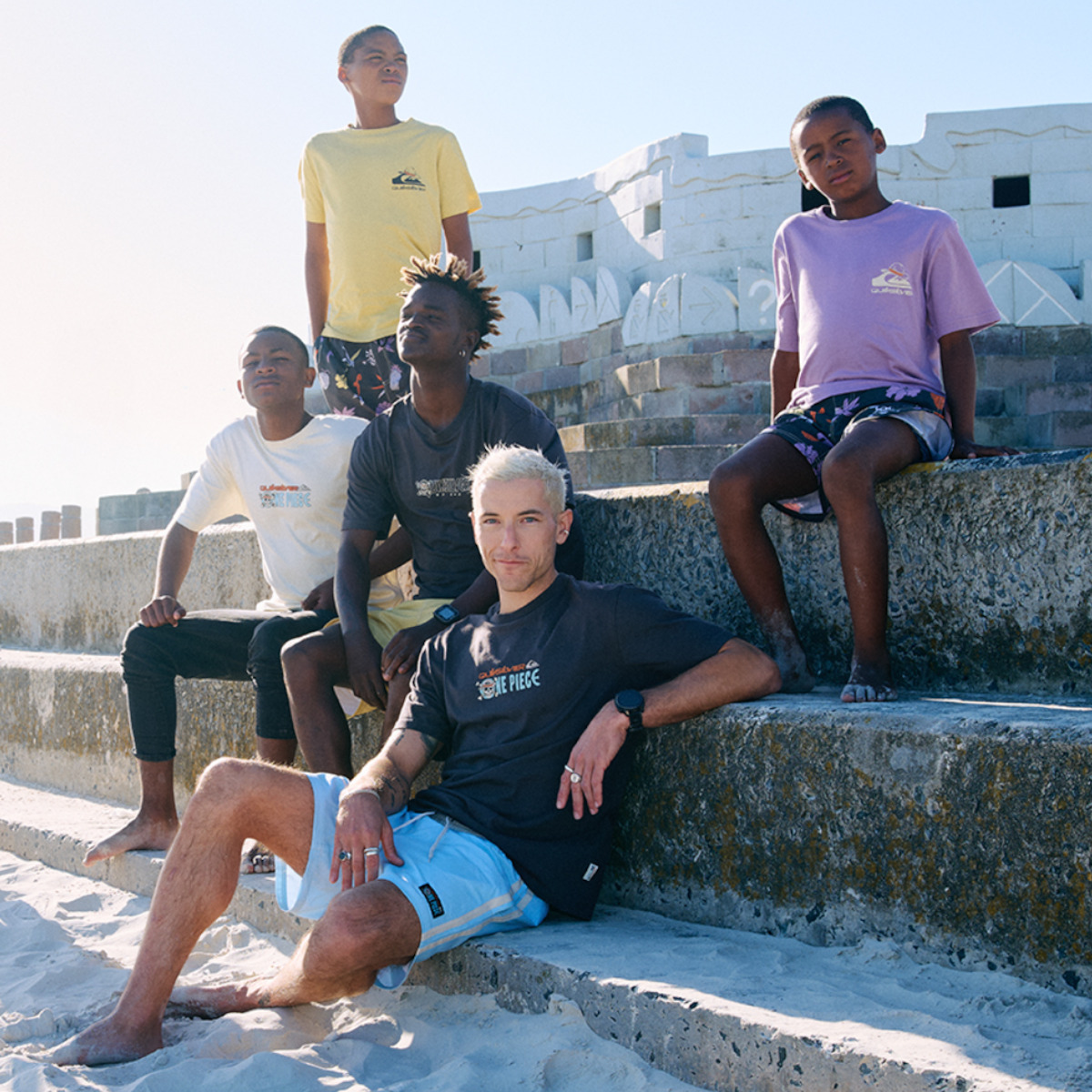 Five people posing on stone steps by the beach, wearing casual t-shirts and shorts, with a bright sky and a large, white building structure in the background. Relaxed, summery atmosphere.