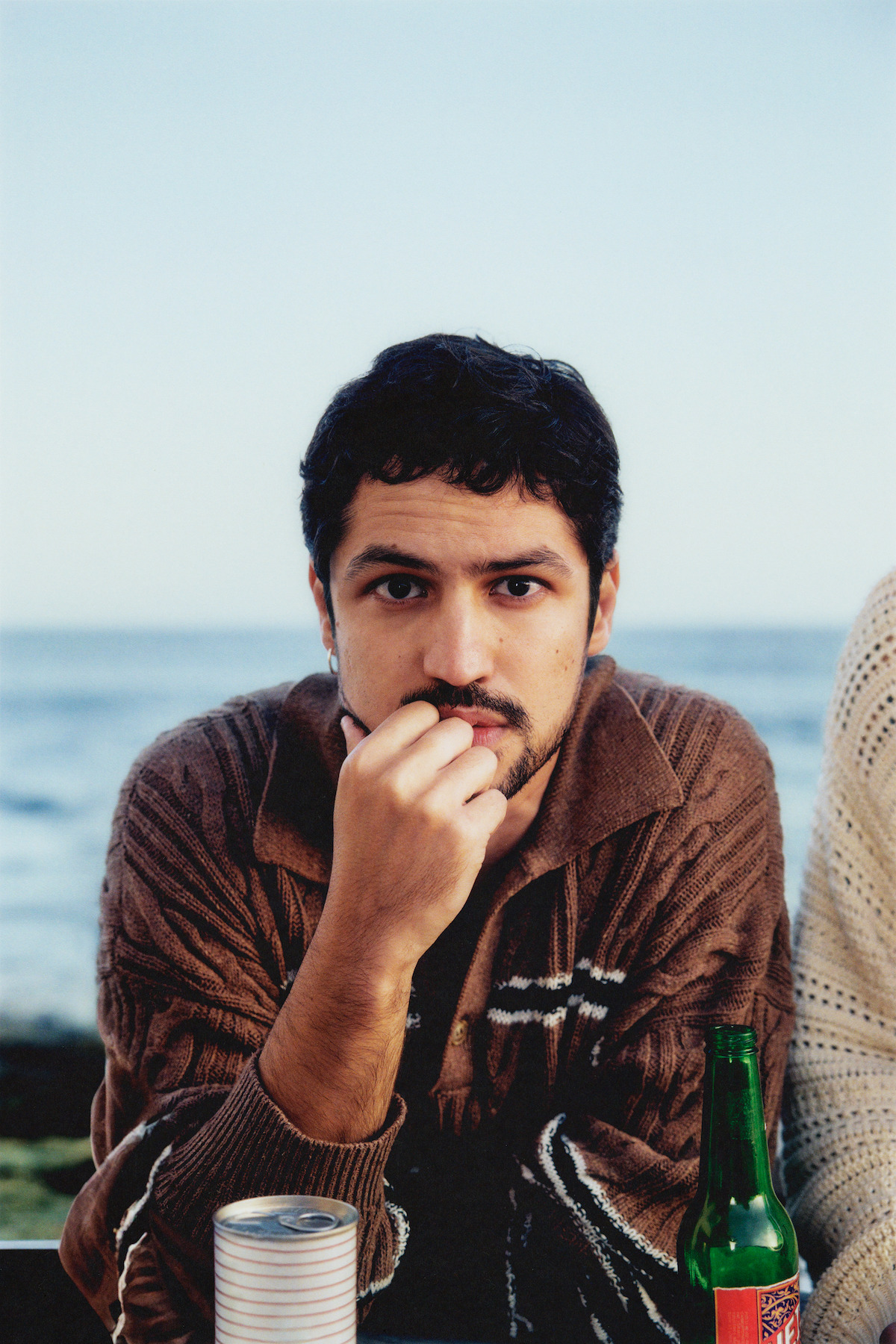 Gabriel Leone wears a brown sweater and sits with a green glass bottle on a beach.