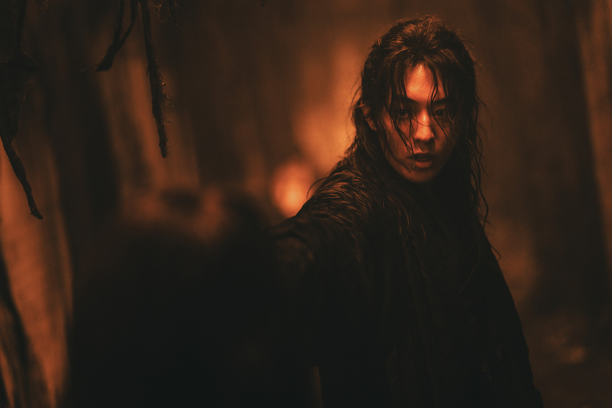 Person with long wet hair in a dark, narrow, dimly lit corridor with an intense expression, reaching toward the camera, dramatic orange lighting creating a suspenseful and mysterious atmosphere.