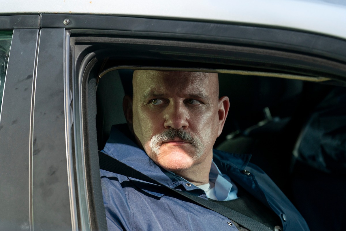 Serious-looking police officer with a mustache sitting inside a patrol car, looking out of the open window, wearing a uniform in a daylight urban environment.