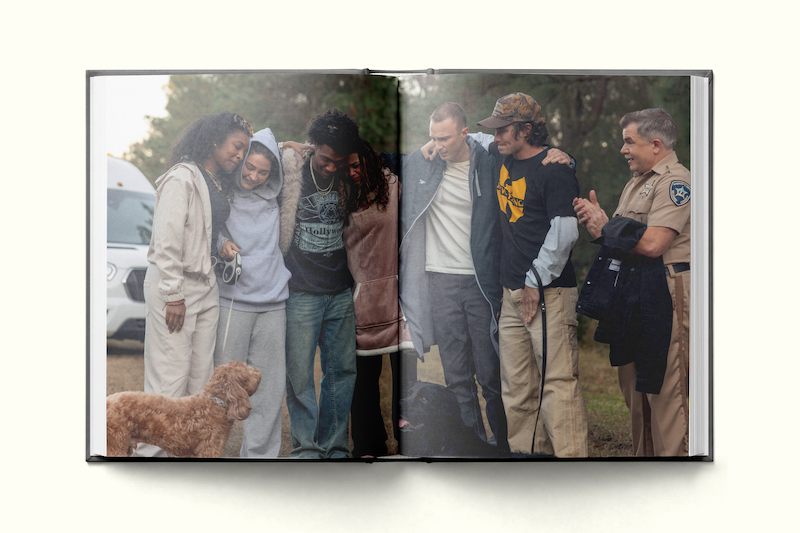 A group of six people in casual clothes stand arm-in-arm outdoors next to a dog, a parked vehicle, and a person in a police uniform, appearing friendly and relaxed in a natural setting.