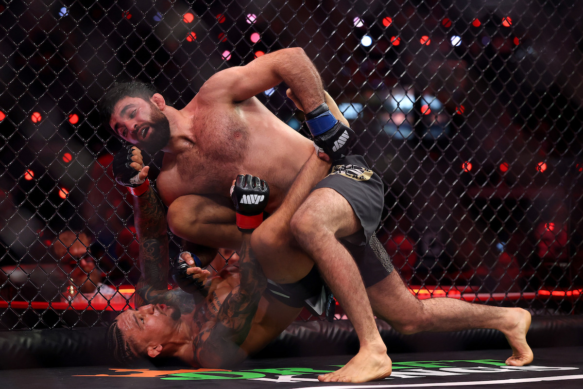 Two male MMA fighters grappling inside a cage during a match, with one fighter on his back attempting a submission hold while the other is on top trying to defend, surrounded by arena lights and a chain-link fence.