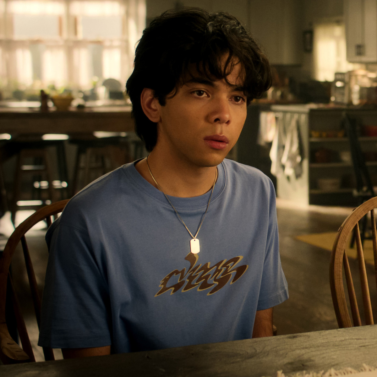 Teen sitting at a wooden table in a cozy kitchen, wearing a blue graphic t-shirt and necklace, looking thoughtful in natural morning light.