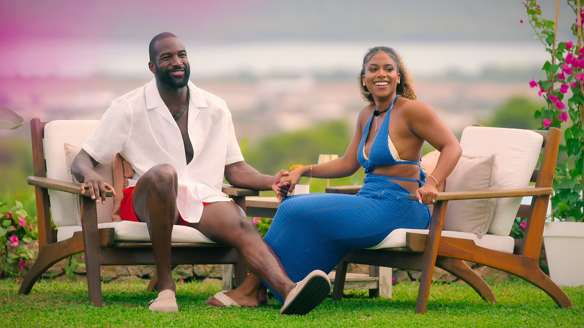 A smiling couple sits on outdoor chairs holding hands in a garden with green grass, flowers, and a scenic blurred landscape in the background, enjoying a sunny day.