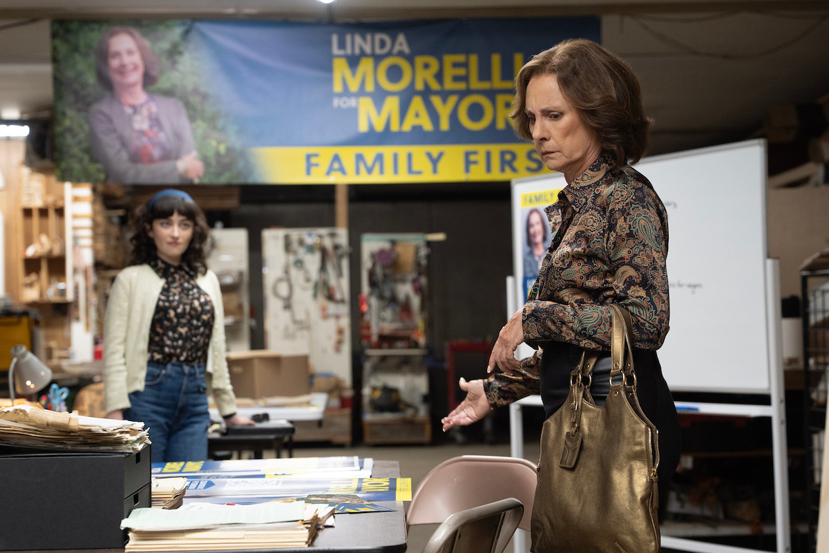 Two women in a campaign office with a "Linda Morelli for Mayor" banner, paperwork, and a whiteboard. One woman stands by a table, the other in the background, suggesting a political campaign planning setting.