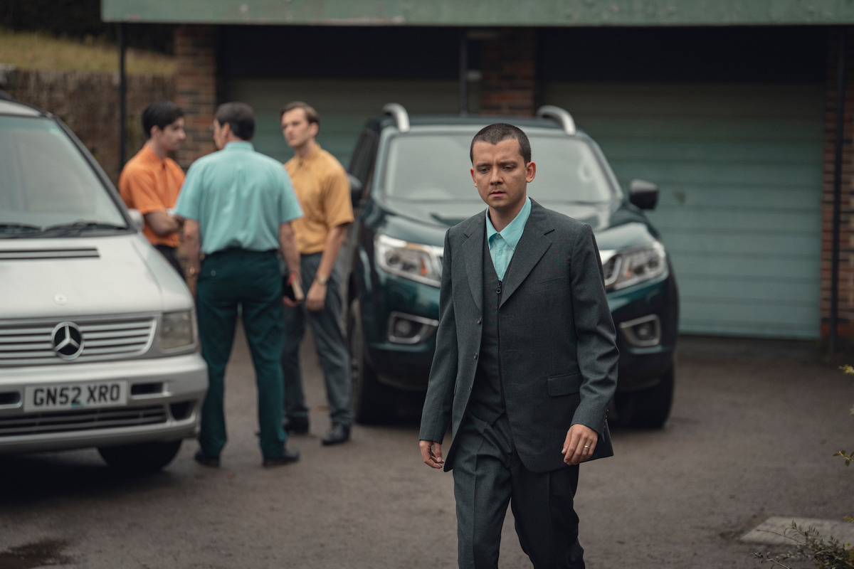 A man in a suit walks in front of two parked cars and a closed garage, while three men stand and talk in the background outside in a driveway.