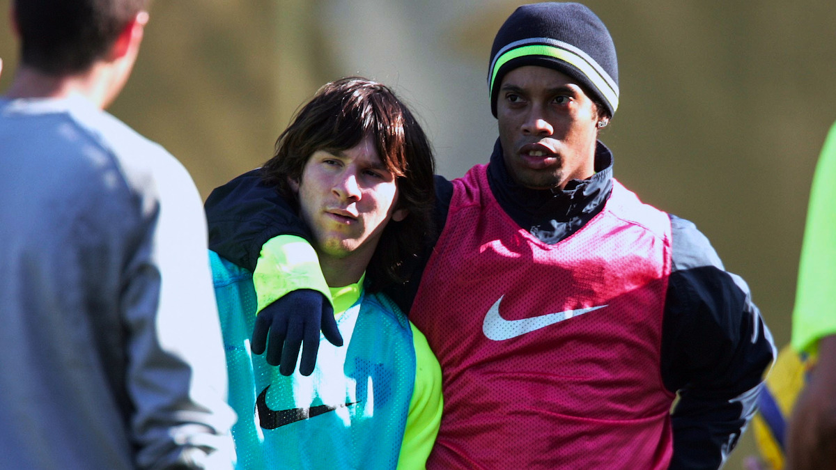 Lionel Messi and Ronaldinho Gaúcho in training bibs stand close together, one with his arm around the other. They are on a practice field, dressed in athletic gear, and appear to be listening to a coach during a team training session.