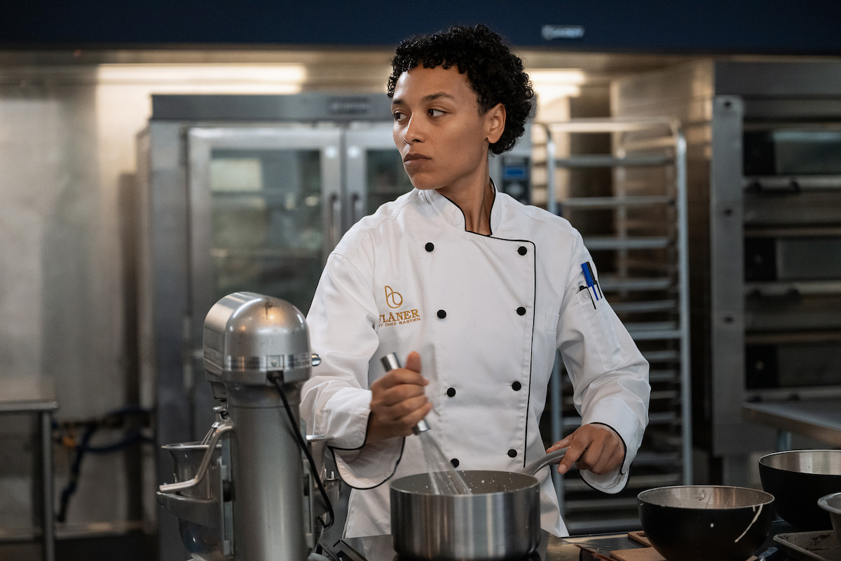 Chef in a white uniform mixing ingredients in a commercial kitchen, surrounded by baking equipment, stainless steel bowls, and industrial ovens in the background.