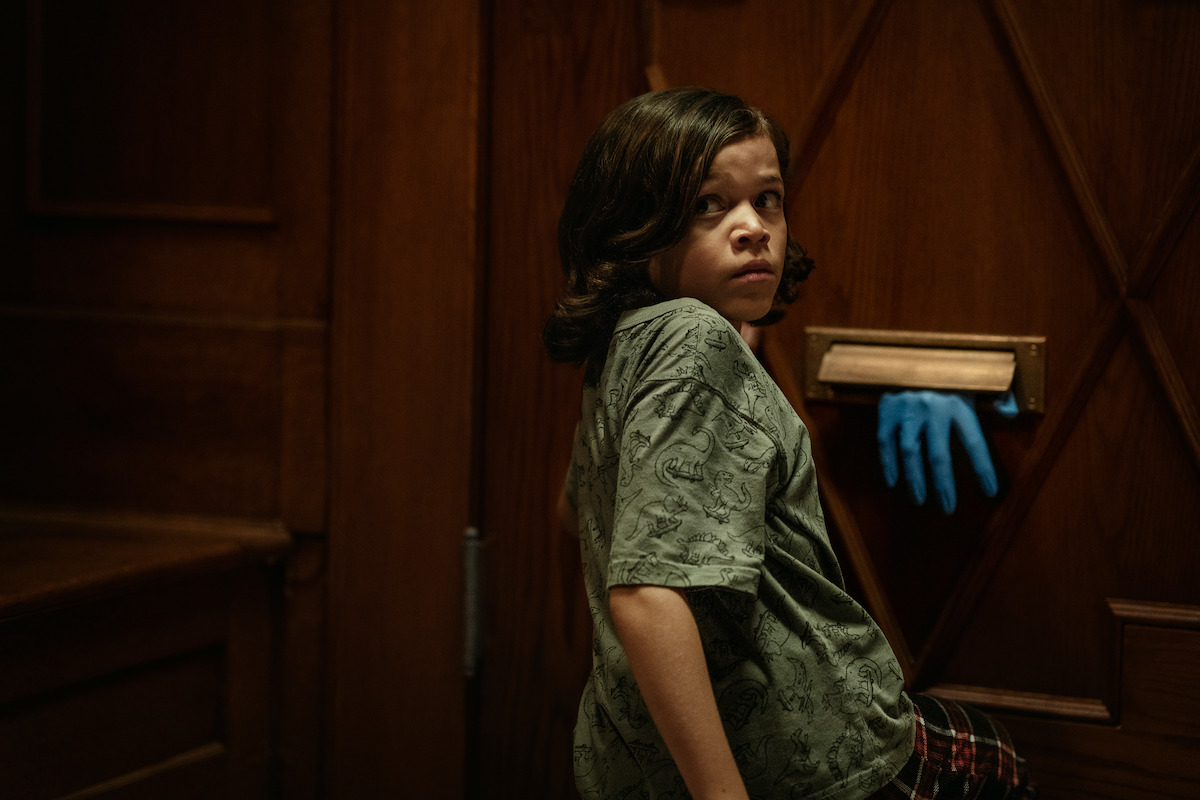 A young person with long hair looks back anxiously in a dimly lit wooden room, standing near a mail slot where a gloved blue hand is reaching through the slot in the door.