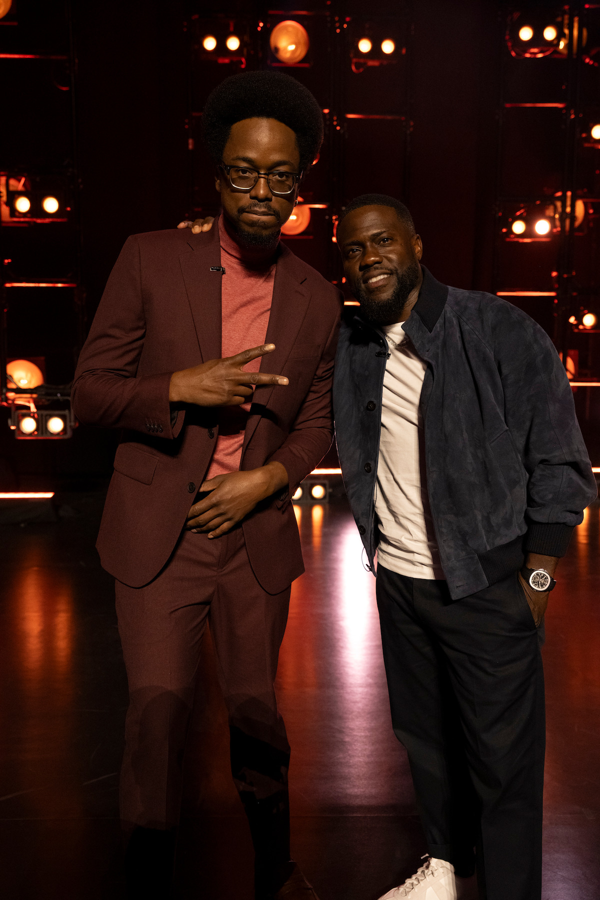 Two men posing together on a stage with dramatic lighting and illuminated bulbs in the background, one wearing a burgundy suit flashing a peace sign, the other in a casual dark jacket and white sneakers, both smiling.