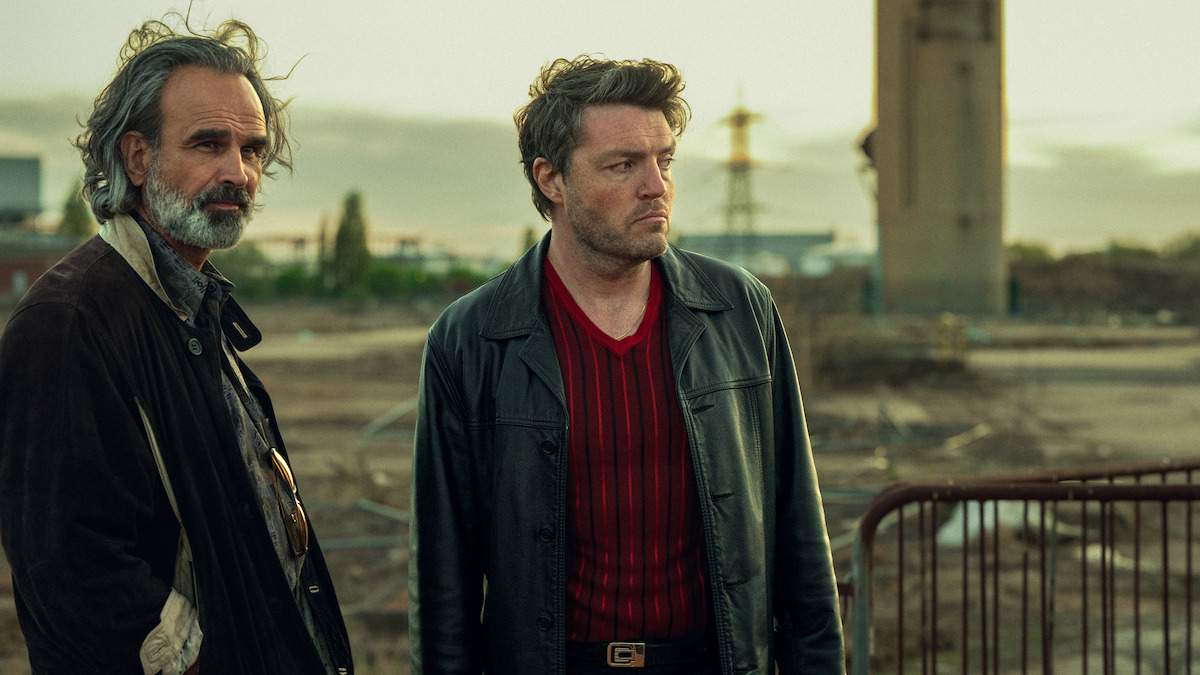 Two serious men stand in an industrial, desolate outdoor setting at dusk, surrounded by dirt, fencing, and distant buildings, creating a tense and moody atmosphere.