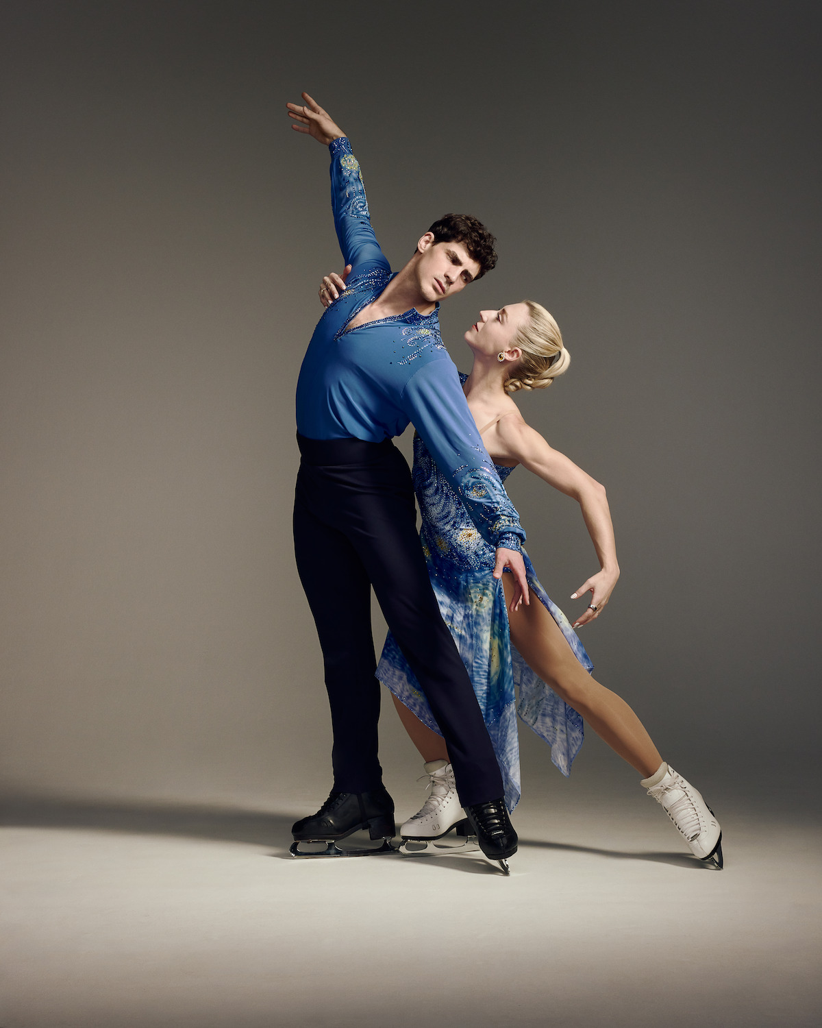 Paul Poirier and Piper Gilles perform an elegant pose on ice in a studio setting, dressed in coordinated blue costumes, with neutral gray background and dramatic lighting highlighting their athleticism and grace.