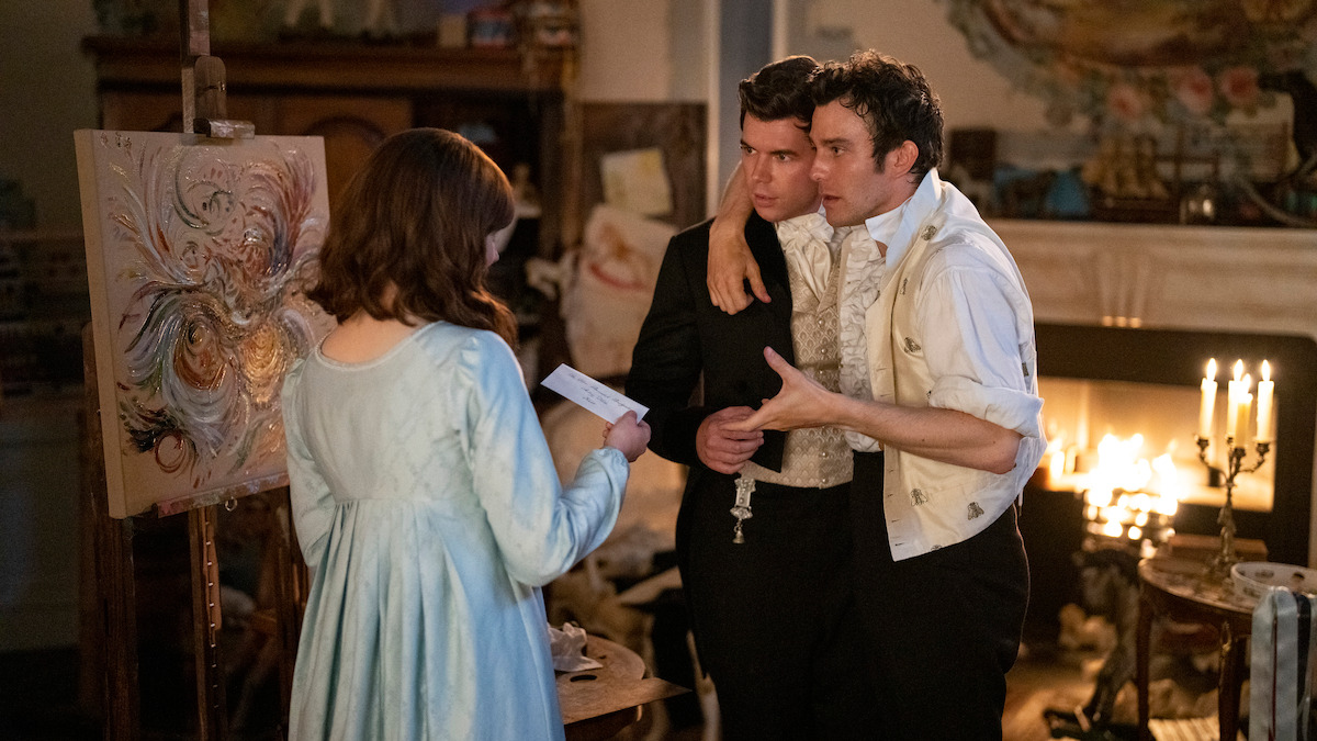 Three people in period clothing stand in a warmly lit, ornate room with a fireplace, candles, and painting supplies; two men talk animatedly while a woman in a blue dress holds a note, suggesting a dramatic or emotional conversation.