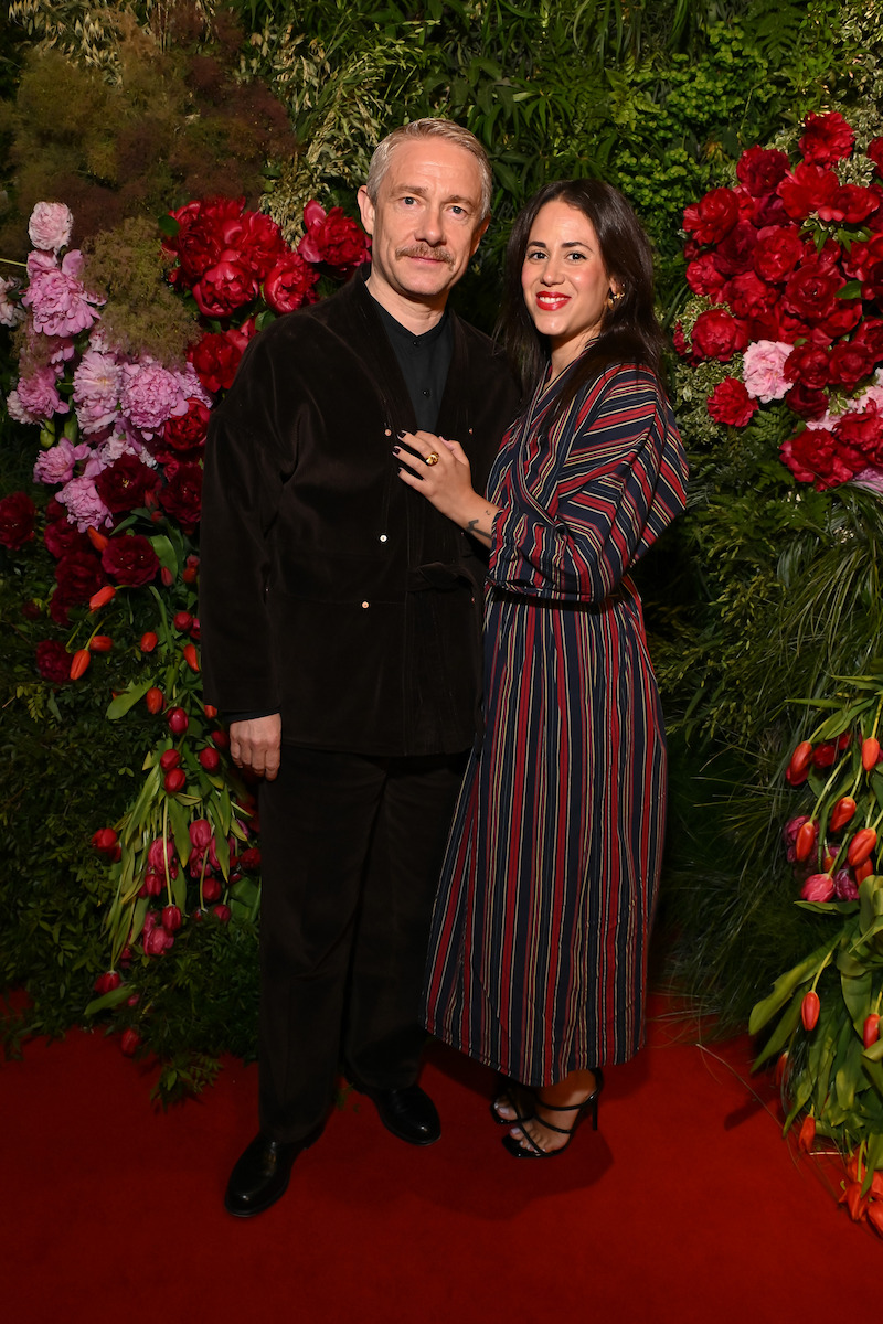 Man and woman posing together on a red carpet, surrounded by lush green foliage and vibrant red and pink flowers, dressed in dark, stylish outfits at an indoor event.
