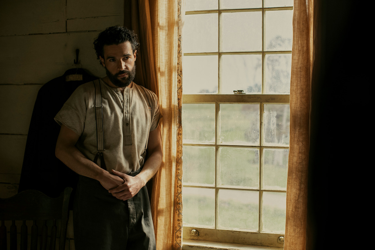 A man with a beard stands pensively near a sunlit window in a rustic room, dressed in an old-fashioned shirt and trousers. The environment suggests a historical or rural setting with muted colors and a calm mood.