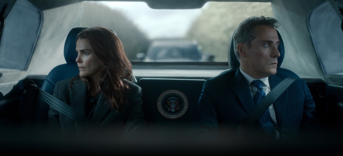 Two serious people in formal attire sit in the back of a car with a presidential seal, looking out the windows; the environment is tense, with another vehicle visible behind them through the rear window.