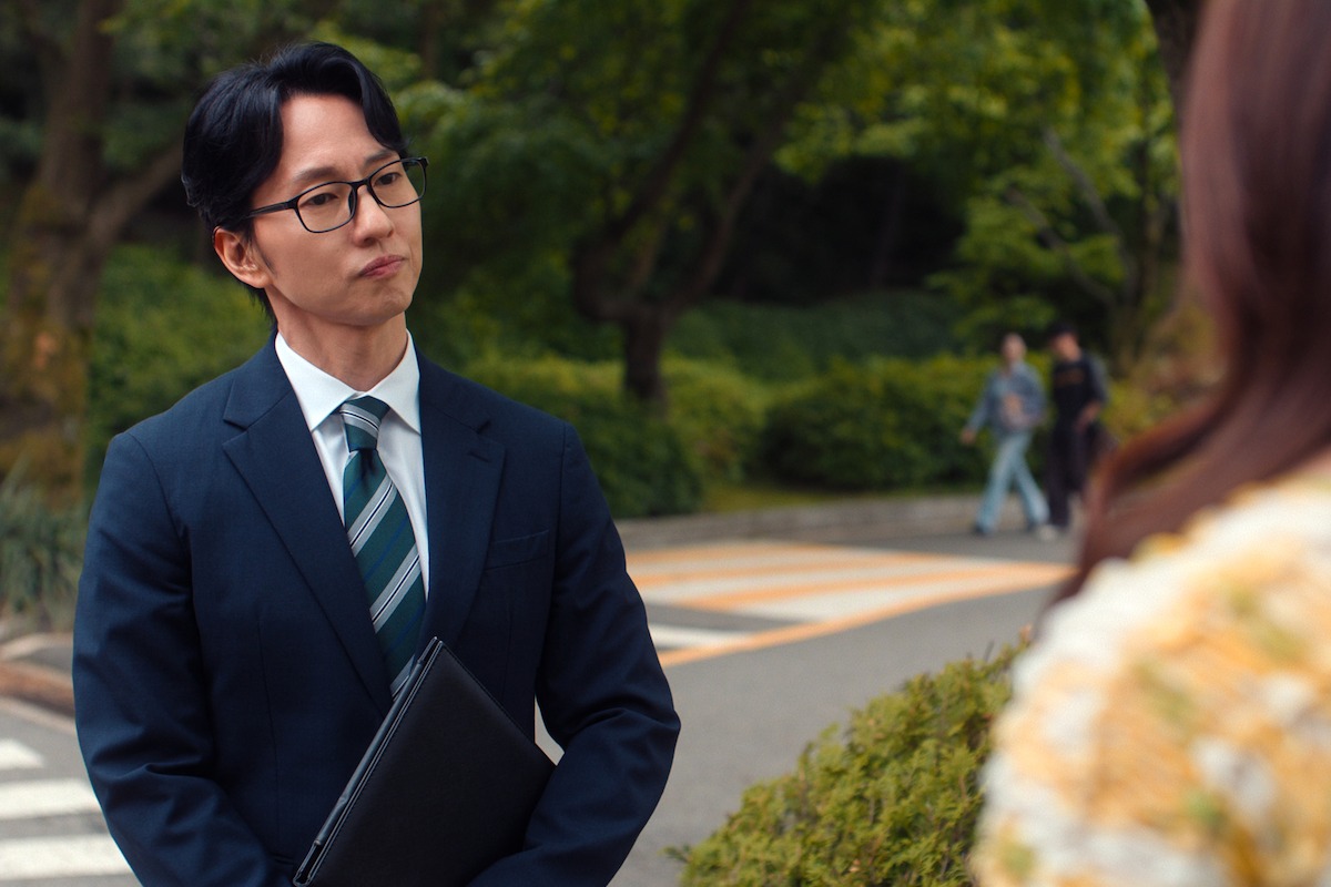Man in suit with glasses holding a folder, standing outdoors on a path in a park with greenery, speaking to a woman in the foreground; two people walking in the background.