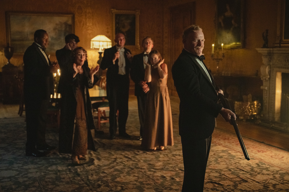 A group of elegantly dressed people in a vintage, dimly lit drawing room; one man in the foreground holding a shotgun, others in the background appear tense or shocked, with classical decor and warm lighting.