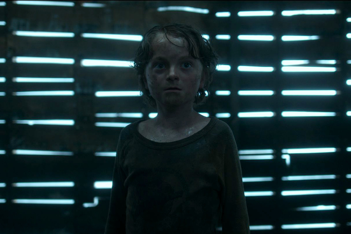 A young, wet child stands in a dimly lit, rustic wooden structure with slatted walls and beams of light shining through the gaps, creating an eerie and suspenseful atmosphere.