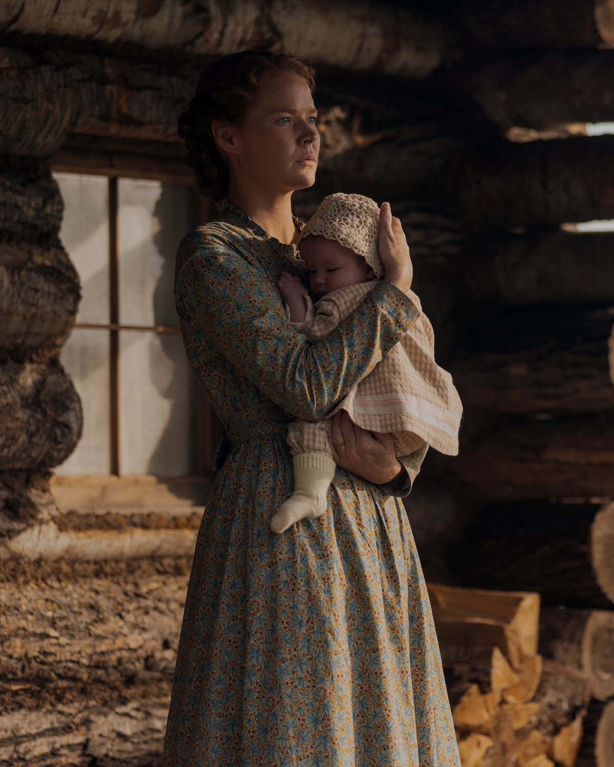 Woman in a floral dress holding a baby outside a rustic log cabin, with sunlight streaming through a window and stacked firewood visible, evoking a historical or pioneer setting.