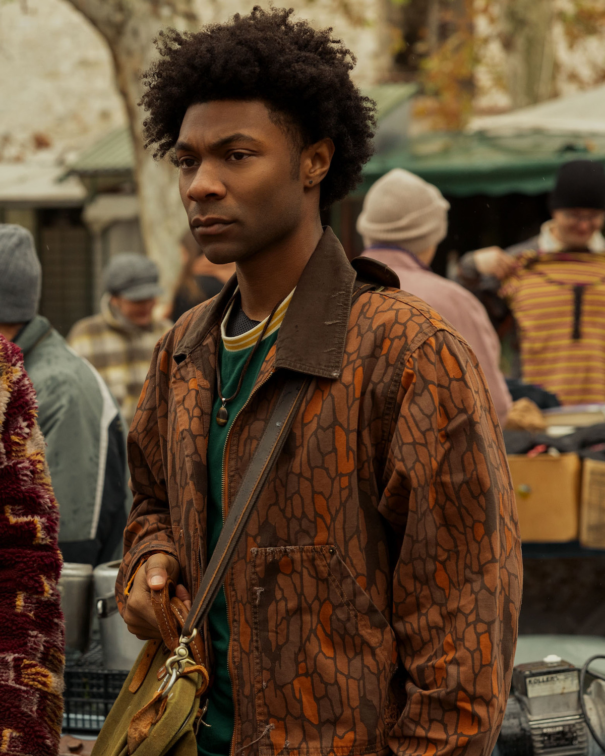 Jonathan Daviss as Pope Heyward, with curly hair wearing a brown patterned jacket and green shirt, carrying a shoulder bag, standing in an outdoor market with blurred people and stalls in the background.