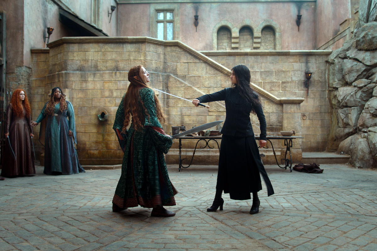 A dramatic sword duel between two women in medieval-style clothing in a stone courtyard, with onlookers watching tensely, evoking a tense and suspenseful atmosphere.