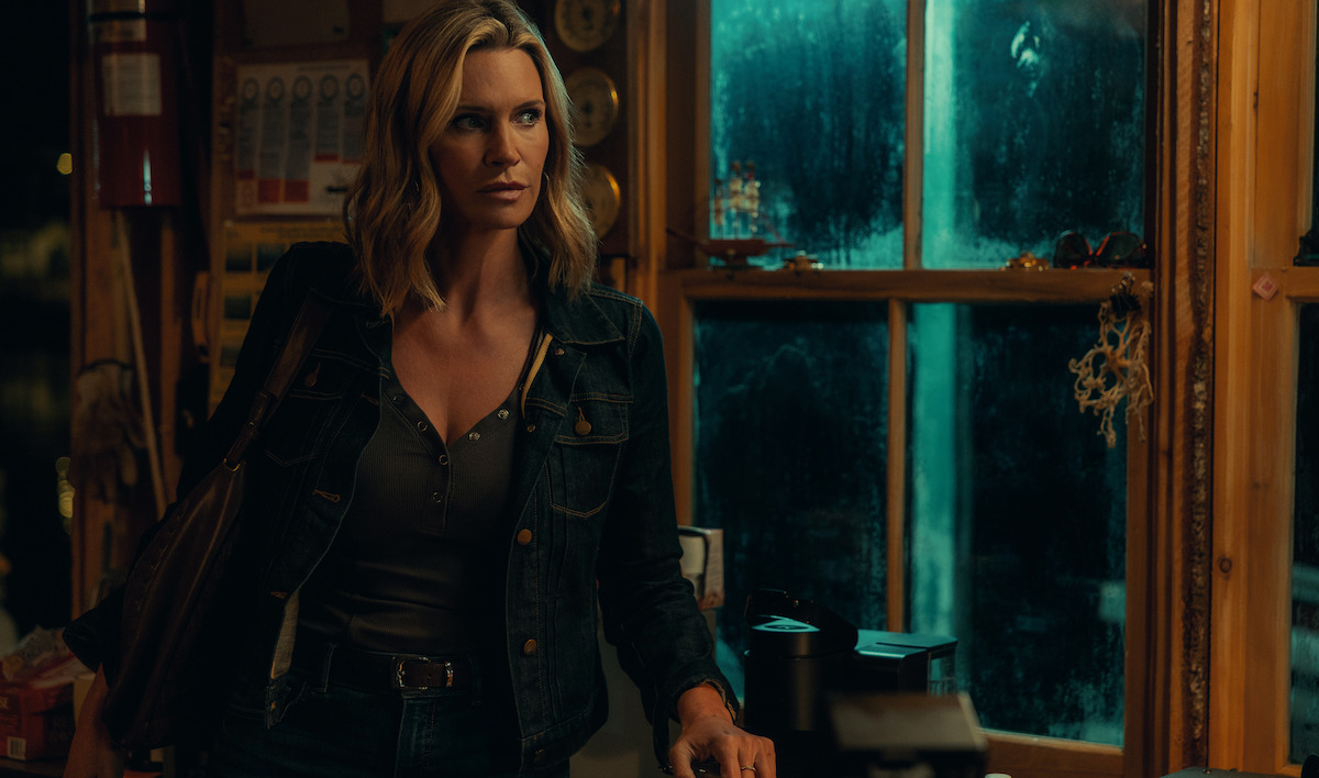A woman with shoulder-length blonde hair stands in a dimly lit room with wooden walls and a foggy window, wearing a dark denim jacket and carrying a bag, looking alert and focused as she faces forward.