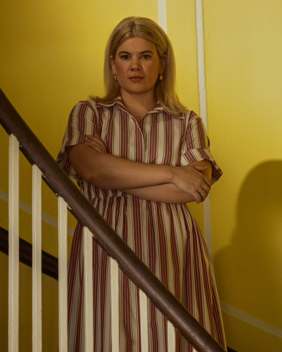Caroline Arapoglou as Rose Cameron, with blond hair in a striped dress stands on a staircase with arms crossed, against a yellow wall with white trim, in indoor lighting.