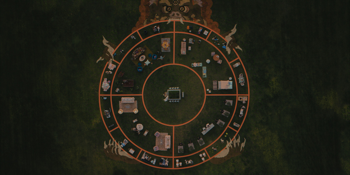 Aerial view of a circular outdoor setup divided into sections with furniture and objects arranged in a pattern on grass, surrounded by artwork of mythical creatures.