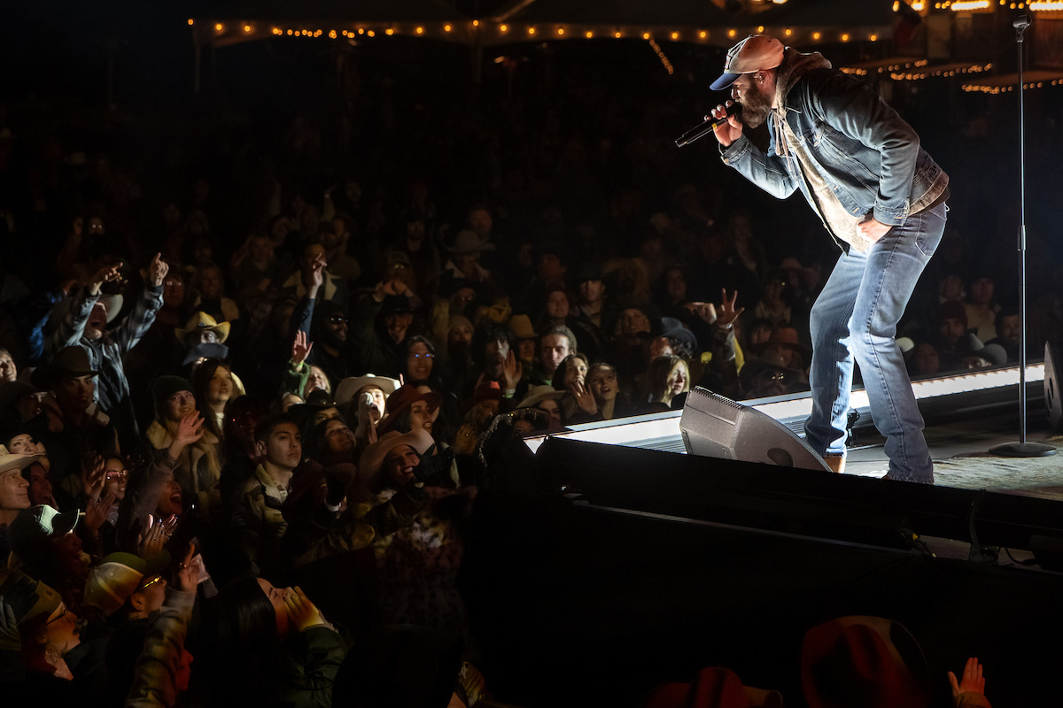 Singer in a hat and denim jacket performing on an outdoor stage at night, illuminated by stage lights, with a large crowd of enthusiastic fans in the foreground and string lights visible in the background.