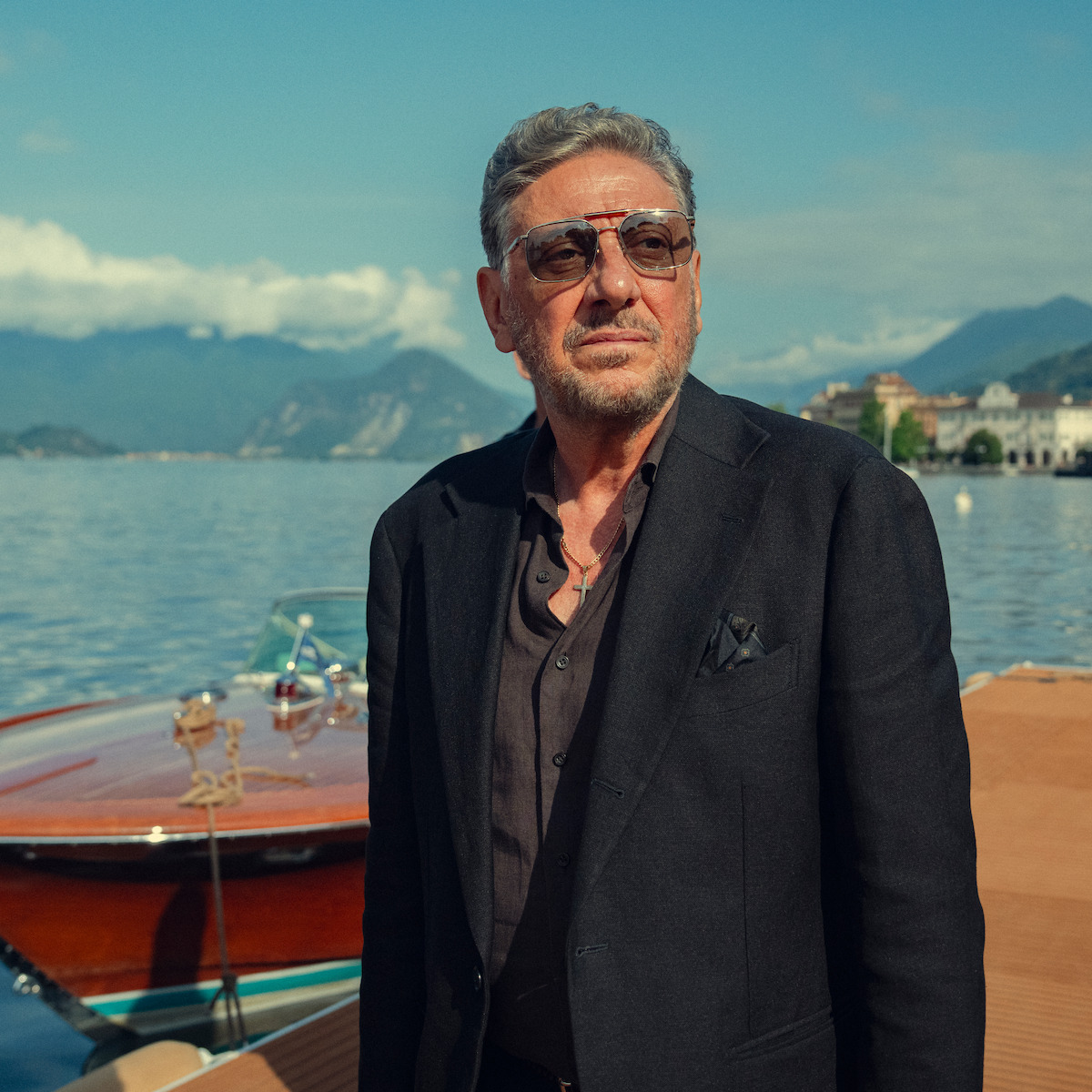 Man in a dark suit and sunglasses stands by a wooden boat on a dock, with a lake, mountains, and town buildings in the background under a clear sky.