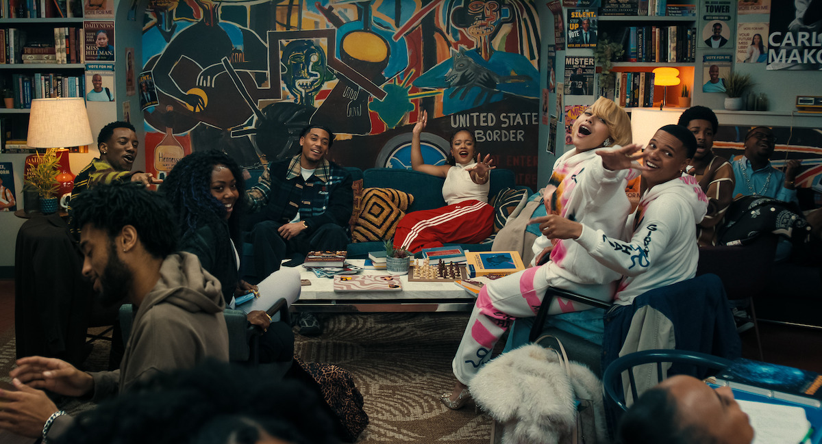 A group of people sit together in a cozy room filled with books and art, laughing and interacting around a coffee table, with colorful murals and shelves in the background, creating a lively and creative atmosphere.