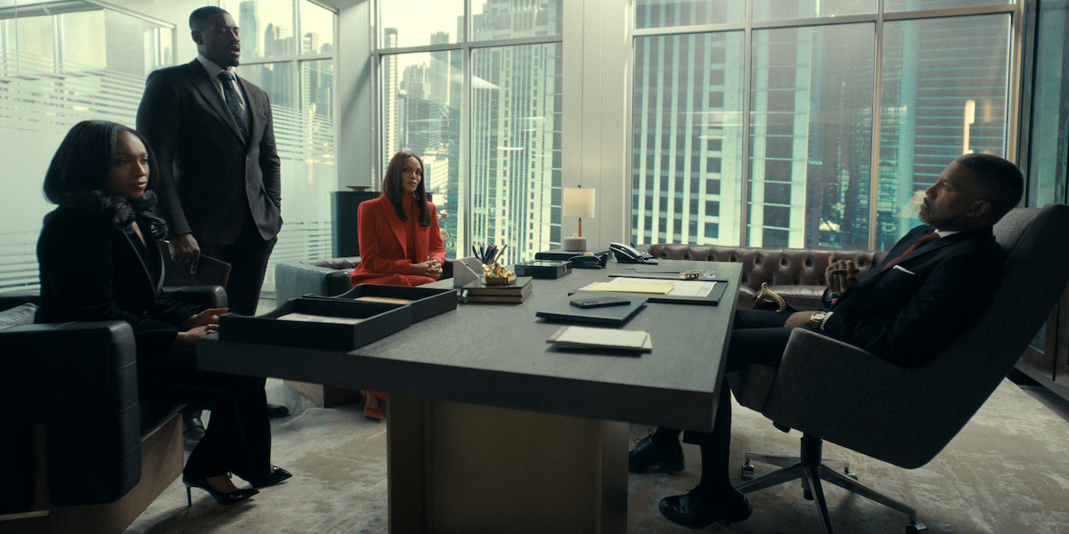 Four professionally dressed people have a serious meeting in a modern office with large windows and city skyscraper views, seated around a large executive desk with paperwork and office supplies.