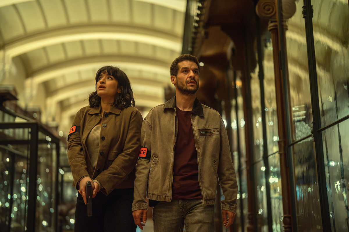 Two people wearing jackets with armbands walk through a dimly lit, ornate hallway lined with glass cases. One holds a gun, both look alert and cautious, as if investigating or searching for something. The setting feels tense and mysterious.