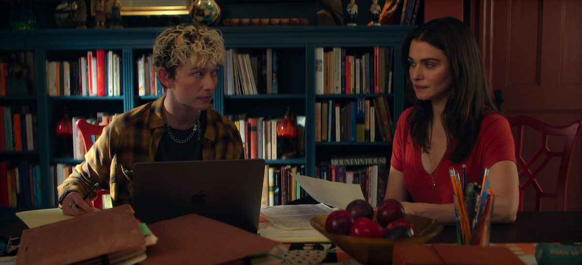 In a cozy library/den lined with bookshelves, Ellen Robertson (Sid) and Rachel Weisz (M) sit at a cluttered table with papers, folders, and laptops, conversing intently. A bowl of apples is in the center of the table.