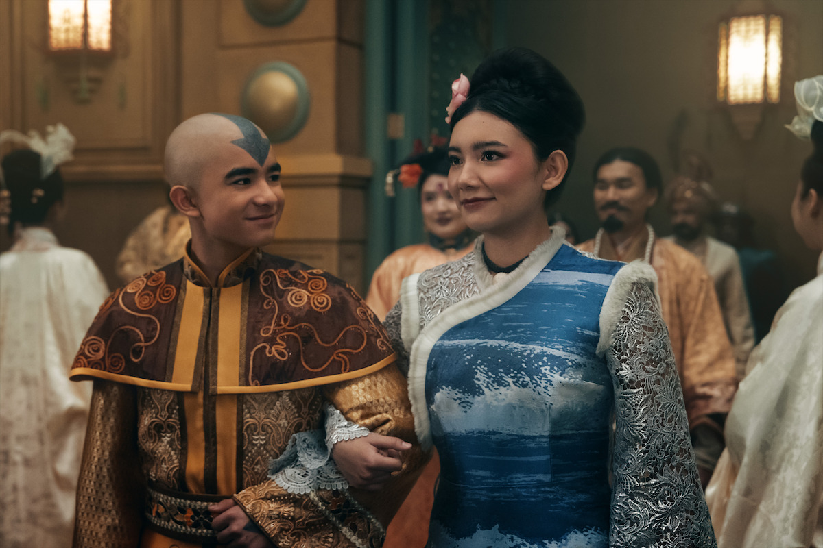 Gordon Cormier as Aang and Kiawentiio as Katarain in elaborate, fantasy-inspired costumes walk arm-in-arm at an elegant indoor gathering, surrounded by others in ornate attire, giving a festive and ceremonial atmosphere.