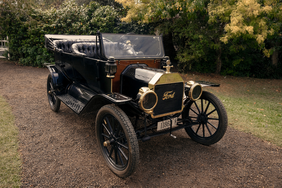 Vintage black Ford Model T parked on a gravel driveway, surrounded by greenery and trees, with a classic open-top design and brass details visible in an outdoor setting.
