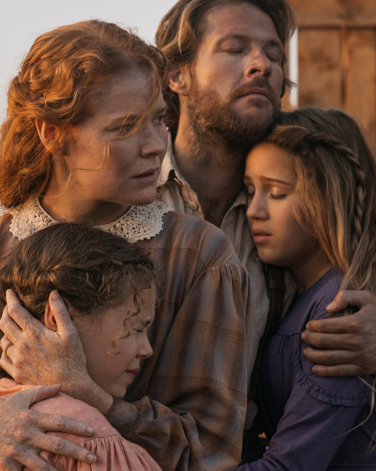 A family of four in old-fashioned clothing embrace closely with worried expressions outdoors, suggesting a historical or frontier setting and a moment of emotional distress or danger.