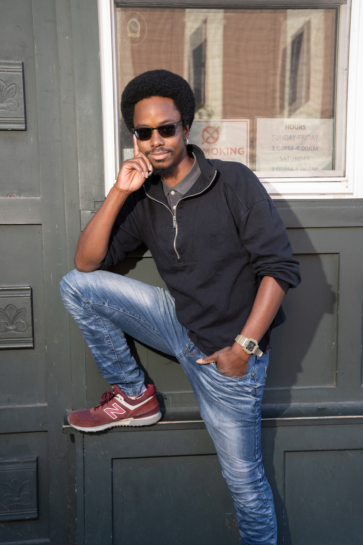 Man wearing sunglasses, black sweatshirt, and jeans poses casually with one leg raised against a green door. A window with signs is behind him, and he is resting his hand on his face, outdoors in an urban setting.