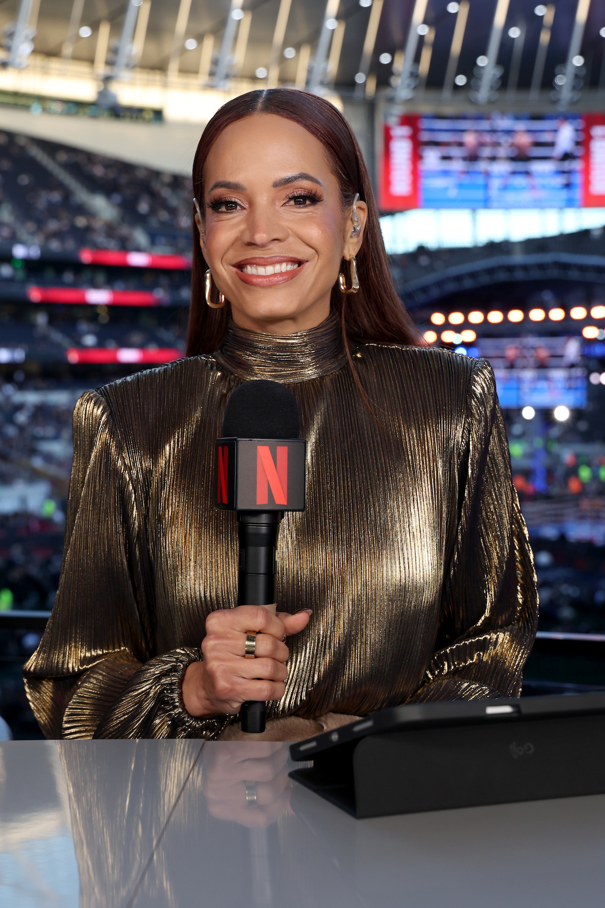Woman in metallic dress holding Netflix microphone, smiling at a desk in a stadium filled with people and bright lights, with a large screen and arched roof structure in the background.